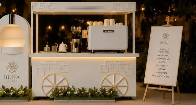 The white BUNA Catering coffee cart lit up at an evening event, with espresso machine, arched lamp panel and menu board